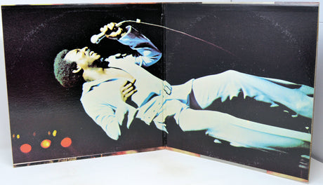 An open vinyl record sleeve or photo album spread showing a performer in a light-colored outfit on a dark stage background, photographed against a white surface.