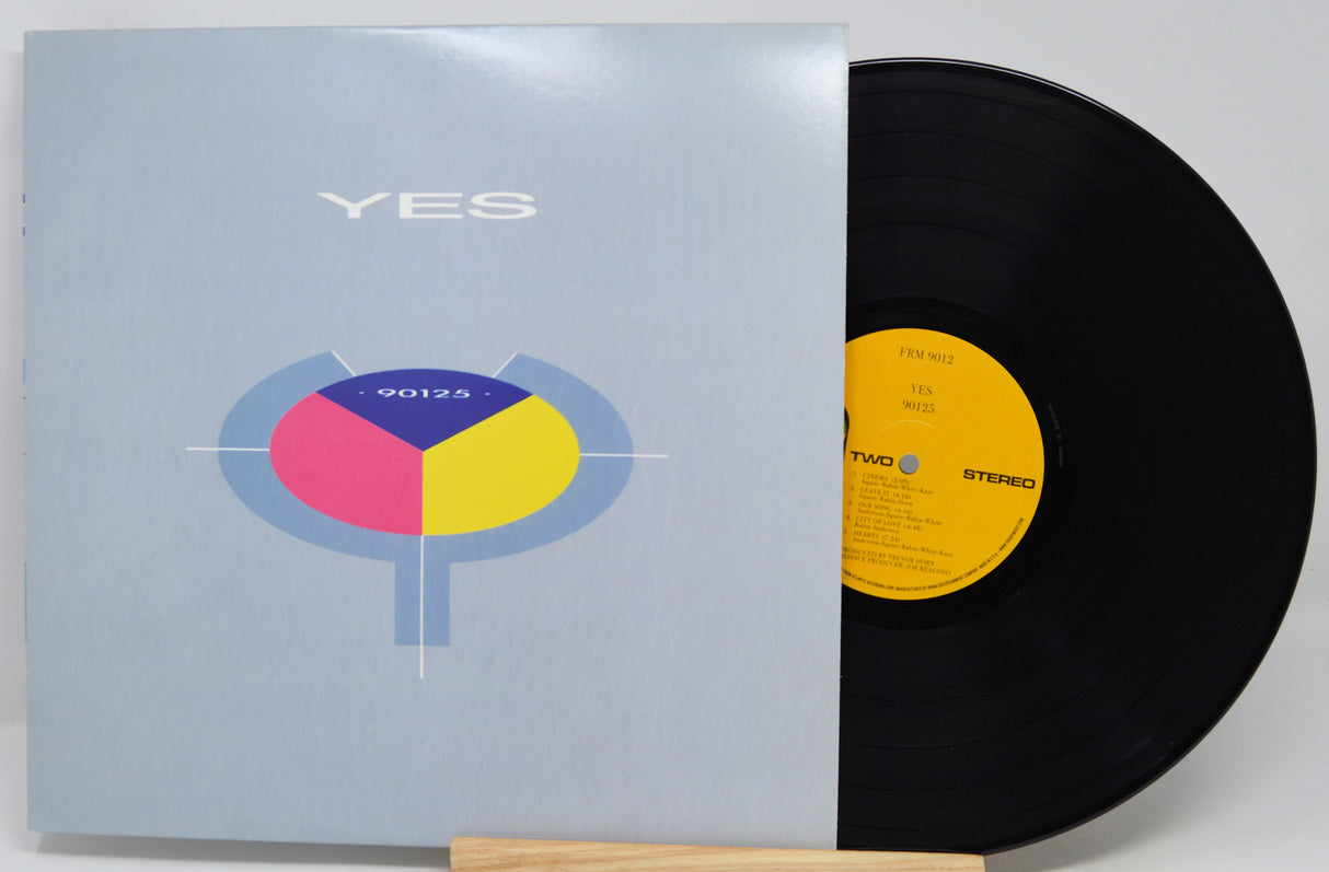 A vinyl record partially pulled from its sleeve, showing the light blue album cover with a colorful circular graphic and the word YES, next to a black record with a yellow label.