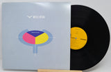 A vinyl record partially pulled from its sleeve, showing the light blue album cover with a colorful circular graphic and the word YES, next to a black record with a yellow label.