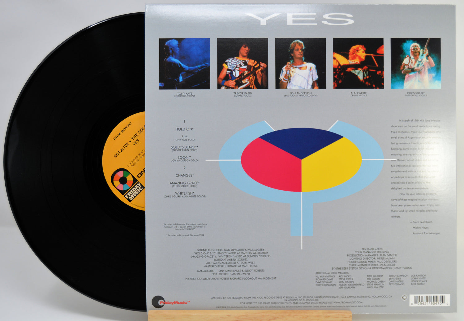 The back of a vinyl record sleeve next to the black record itself, showing the tracklist, artist photos, and a large circular graphic with red, yellow, and blue sections on a light gray background.