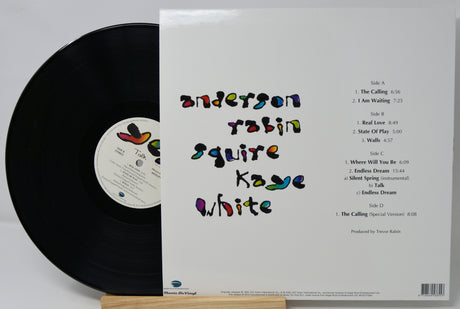 Vinyl record album shown with the black record partially visible on the left and the white outer sleeve on the right, displayed upright on a small wooden stand against a light gray background.