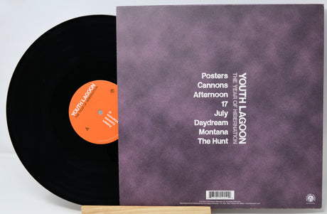 A vinyl record partially pulled from its sleeve, showing a black disc with an orange label next to a mauve-purple cover printed with tracklist text and the band name YOUTH LAGOON.