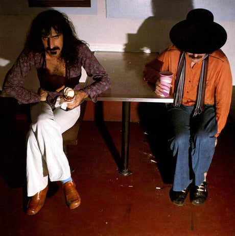 Two men sitting at a table in a casual setting, one holding a cup.