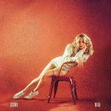 The image features a vinyl record cover with a woman seated on a chair, leaning forward with her hands resting on her knees. She is wearing a white top and white shoes, with her hair styled in a casual, flowing manner.