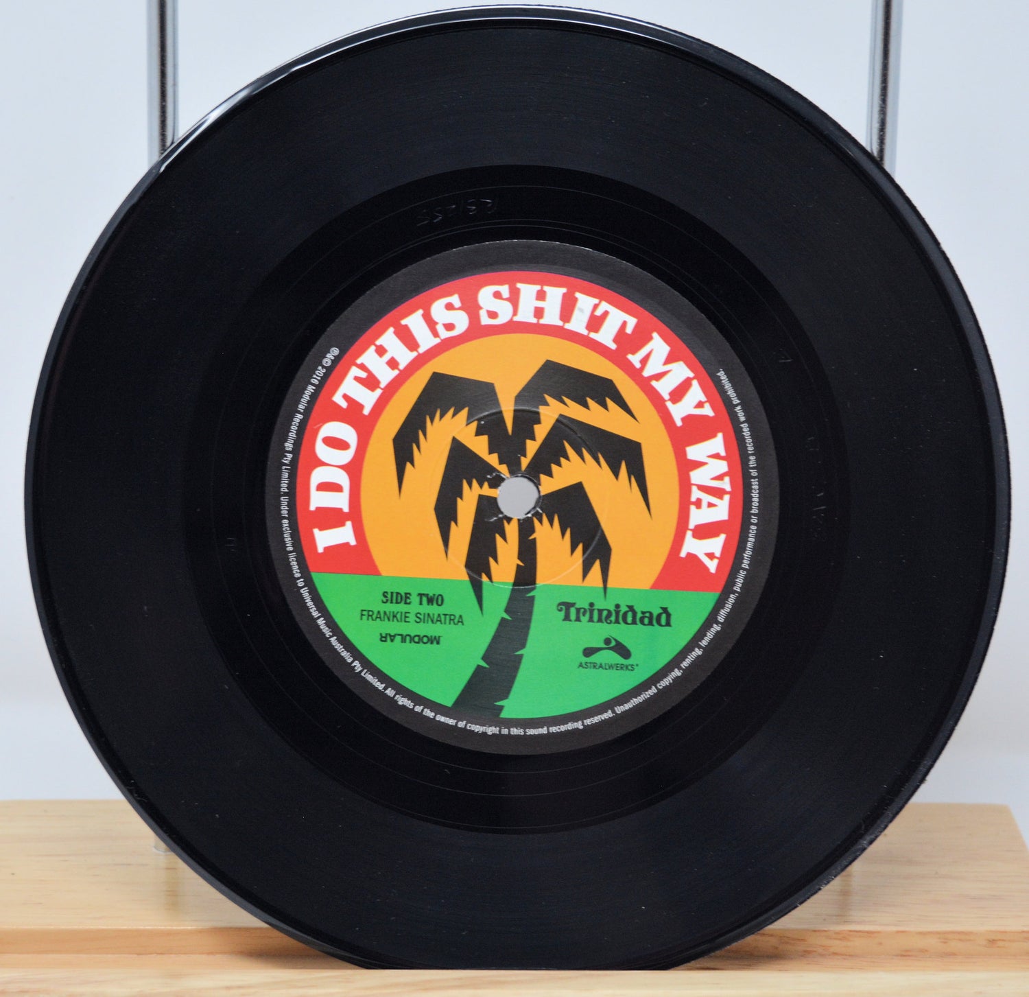 A 7-inch record with a label featuring text and a palm tree graphic. The record is black, with a red and yellow label.