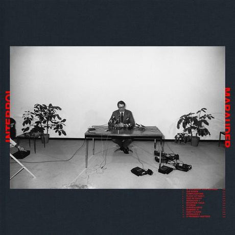 Black and white album cover featuring a person sitting at a desk with a microphone, flanked by two potted plants.