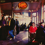 Album cover of 'Muswell Hillbillies' by The Kinks featuring an indoor scene with people sitting and standing, with a prominent sign overhead.