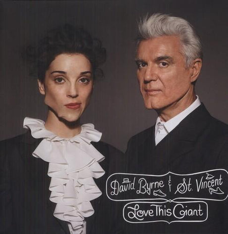 An album cover featuring two individuals, with one wearing a white ruffled shirt and the other in a black suit with white hair. The text on the cover reads 'David Byrne & St Vincent Love This Giant'.