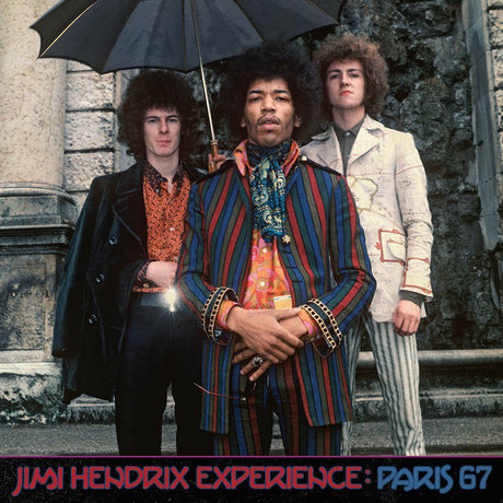 Jimi Hendrix Experience members with Jimi Hendrix in the center, holding an umbrella, against a stone wall background.
