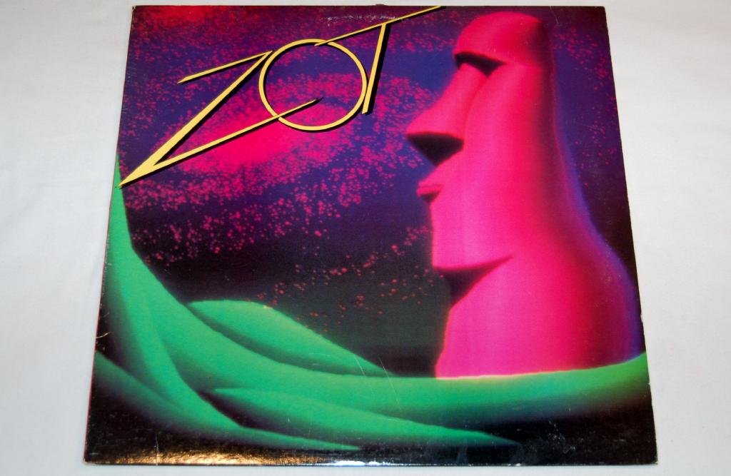 This image features the album cover for "Zot - Zot," a vinyl record. The artwork is striking and abstract, portraying a vibrant pink sculpture of a human face with an oversized nose, set against a dark, textured background.