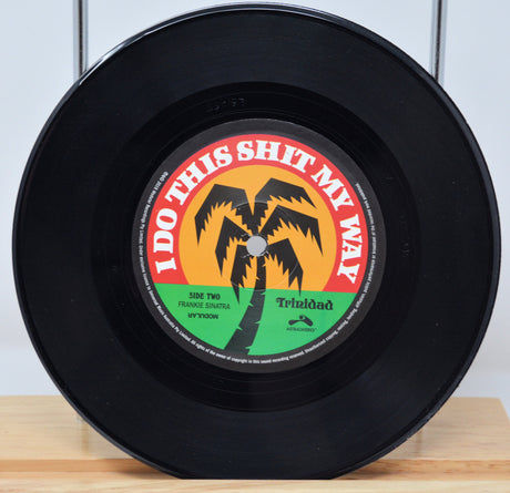 A 7-inch record with a label featuring text and a palm tree graphic. The record is black, with a red and yellow label.